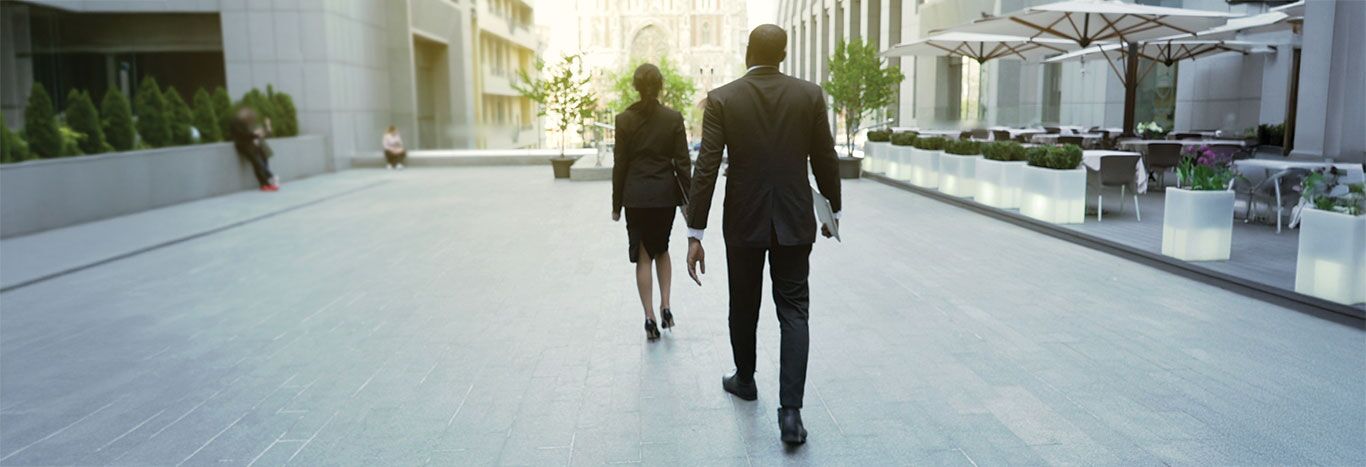 A professionally dressed man and woman walk away down a modern, upscale urban plaza surrounded by tall office buildings.
